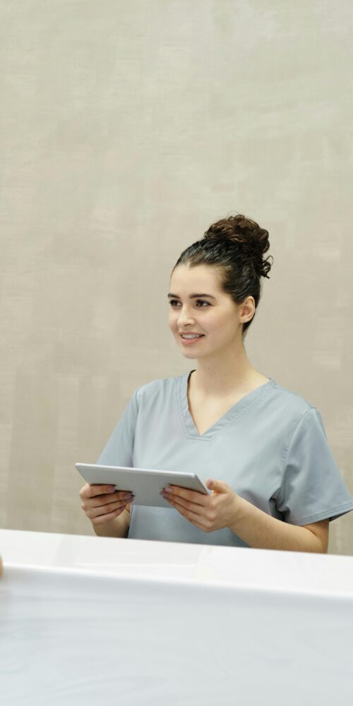 A female medical receptionist in scrubs is smiling and holding a tablet while assisting a woman at a clinic front desk.