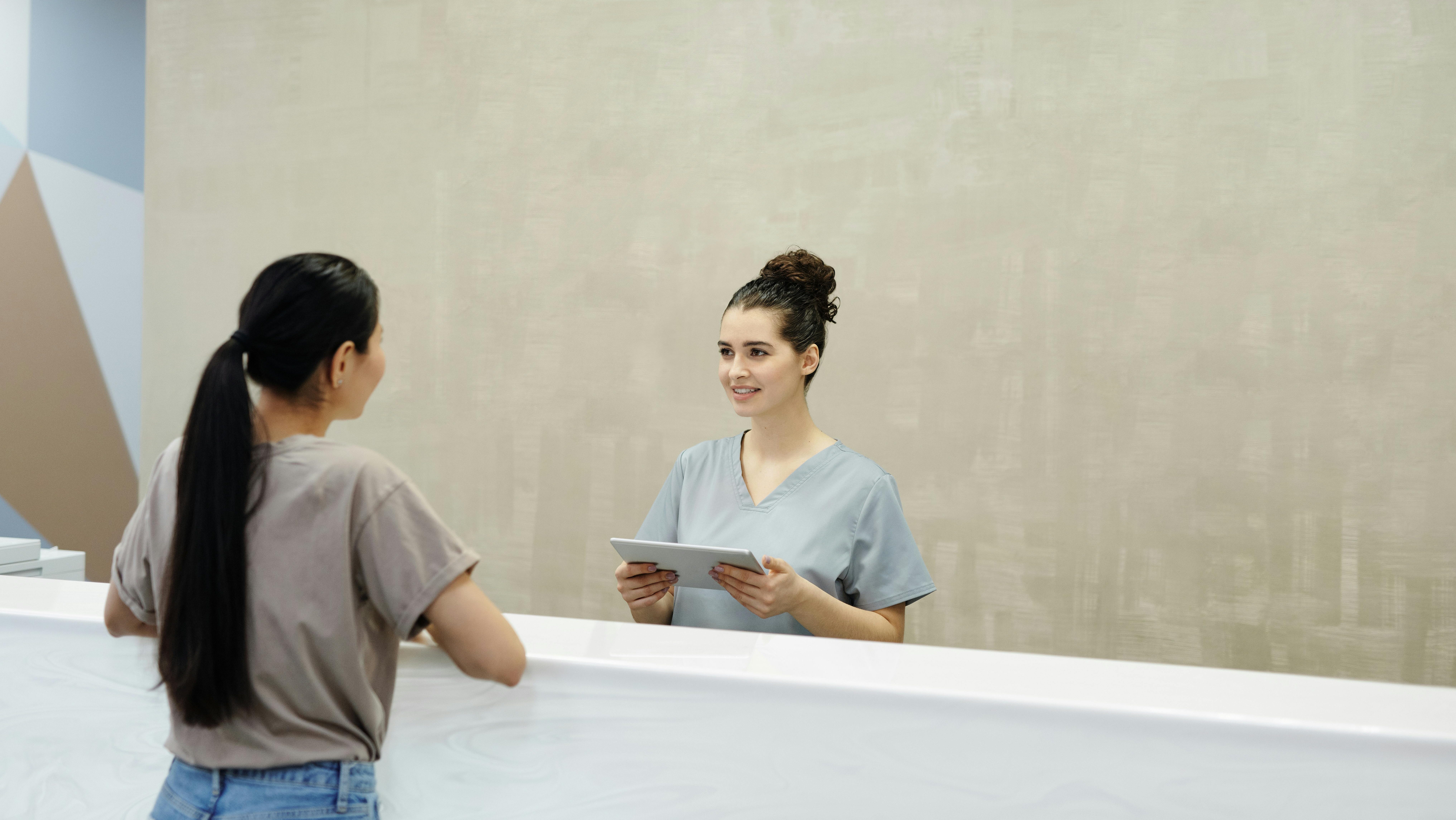 A female medical receptionist in scrubs is smiling and holding a tablet while assisting a woman at a clinic front desk.
